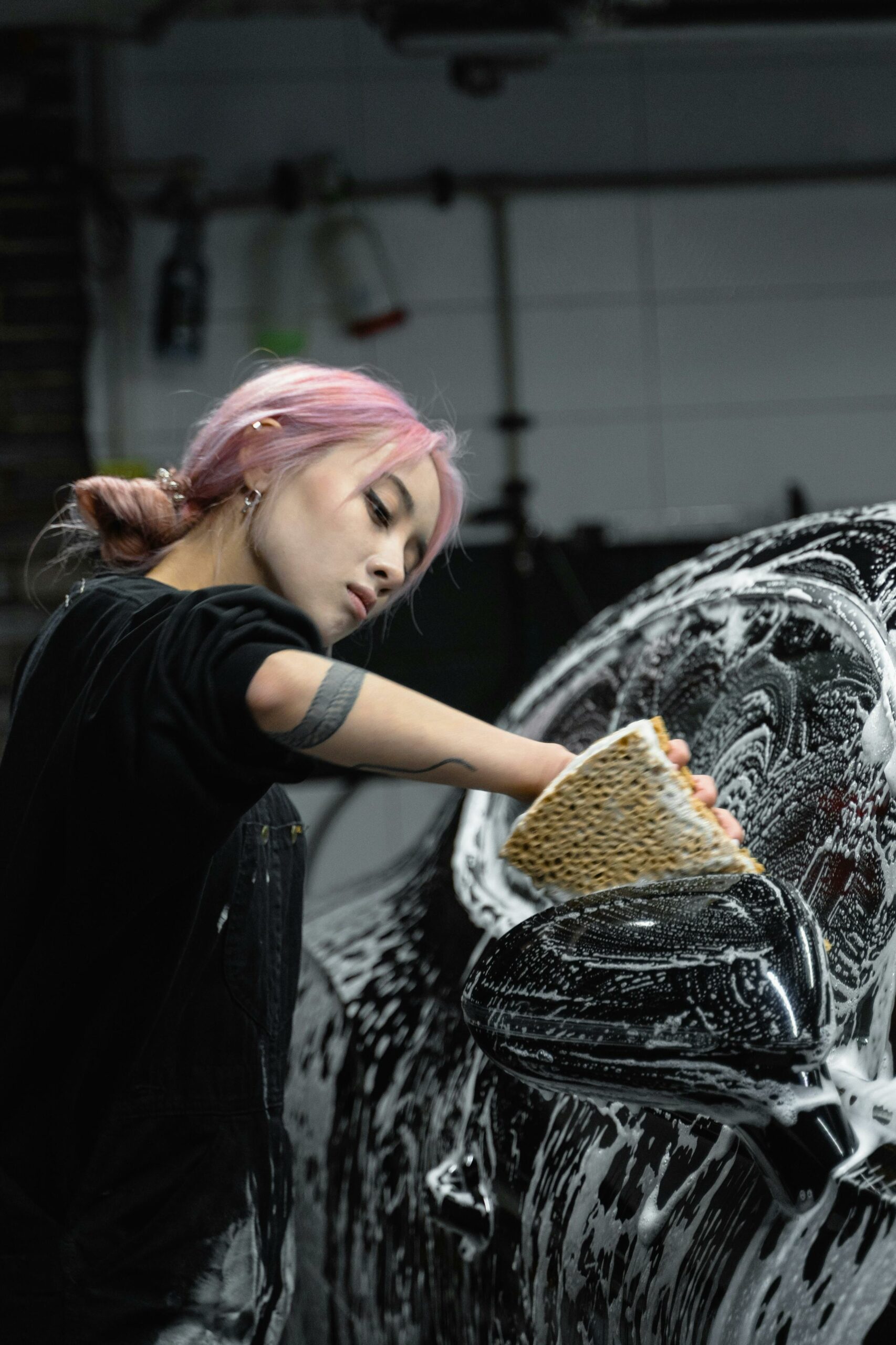A young woman with dyed hair and tattoos cleaning a car with soap and sponge in a garage.